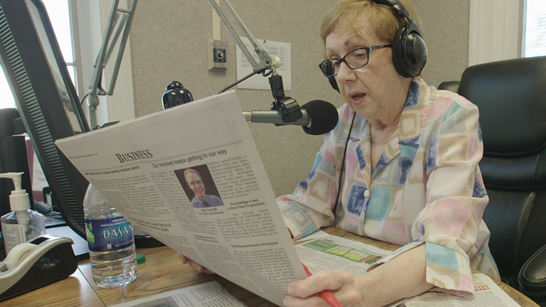 A woman reads a newspaper into a microphone