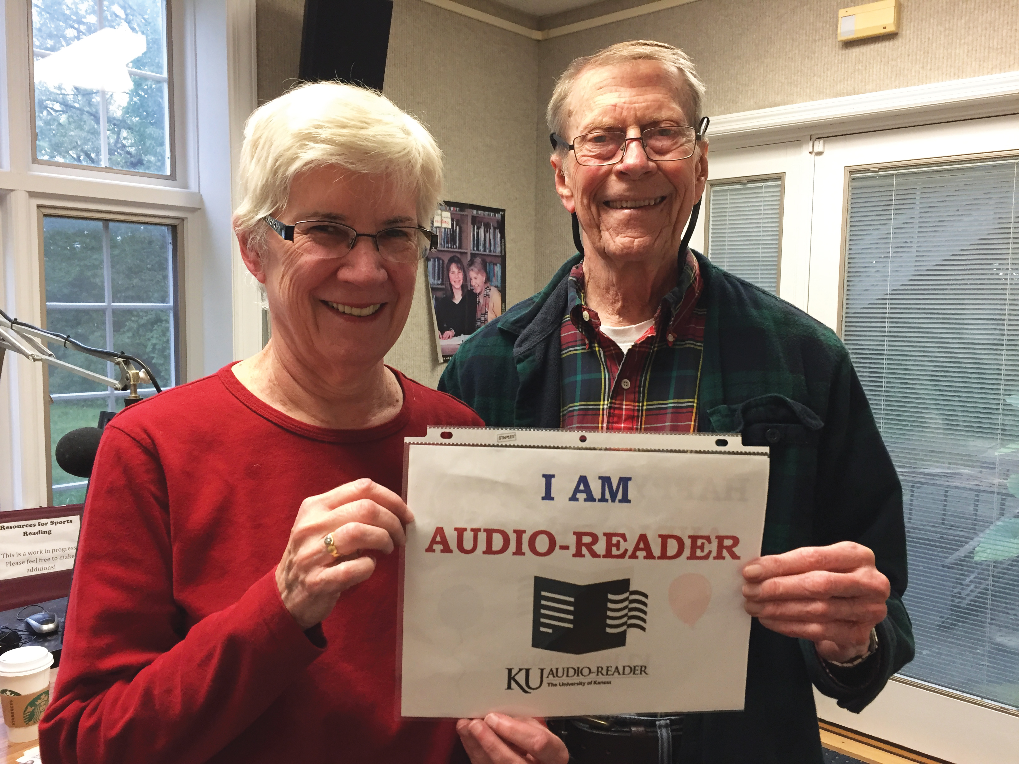 Elinor stands to the left with short grey hair wearing a red shirt. Harlan stands to the right wearing a plaid jacket. Together they hold a sign that reads "I am Audio-Reader"