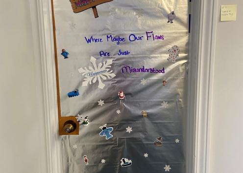 Door number 11 is wrapped in silver wrapping paper and features characters from the 1964 TV special 'Rudolph the Red-Nosed Reindeer.' Written on the silver paper, it says "Where Maybe Our Flaws are Just Misunderstood." There are two white cut-out snowflakes at the bottom of the door and one in the middle that says "uniqueness." 