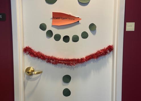 Door number seven is decorated to look like a snowman. The all-white door has three black circles for buttons, red garland for a scarf, black circles for eyes and a smile and an orange, sideways triangle for the nose. 