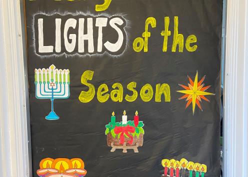 Door number 8 says features a black background with yellow letters that say "Many are the lights of the season." It includes images of a Hanukkah Menorah, an Advent Wreath, a Kwanzaa Kinara to represent all lights of the season.