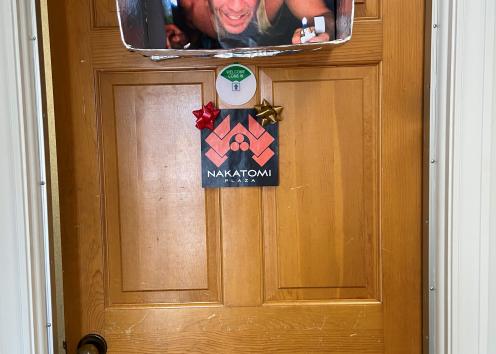 Door number 9 features a 'Die Hard' theme. At the top of the door is a cardboard box wrapped in foil with a picture of Bruce Willis, representing an image from the movie. It has a sign underneath the cardboard box that says 'Nakatomi Plaza.'